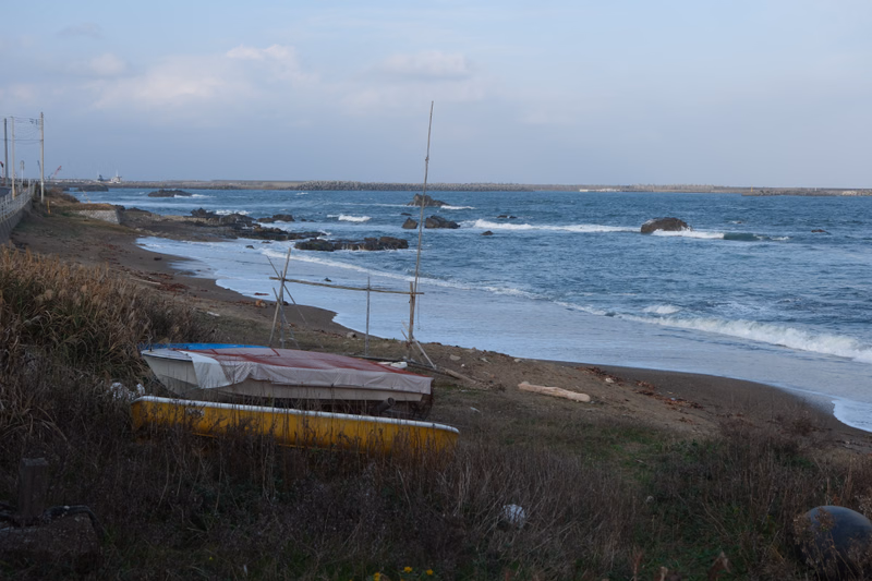 A view of a sandy beach with several beach huts and rocks along the coastline under a pale sky.