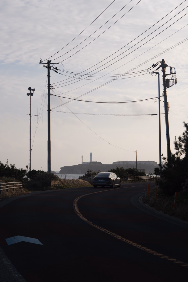 A winding road leads toward a distant coastline under a cloudy sky, with numerous power lines overhead.