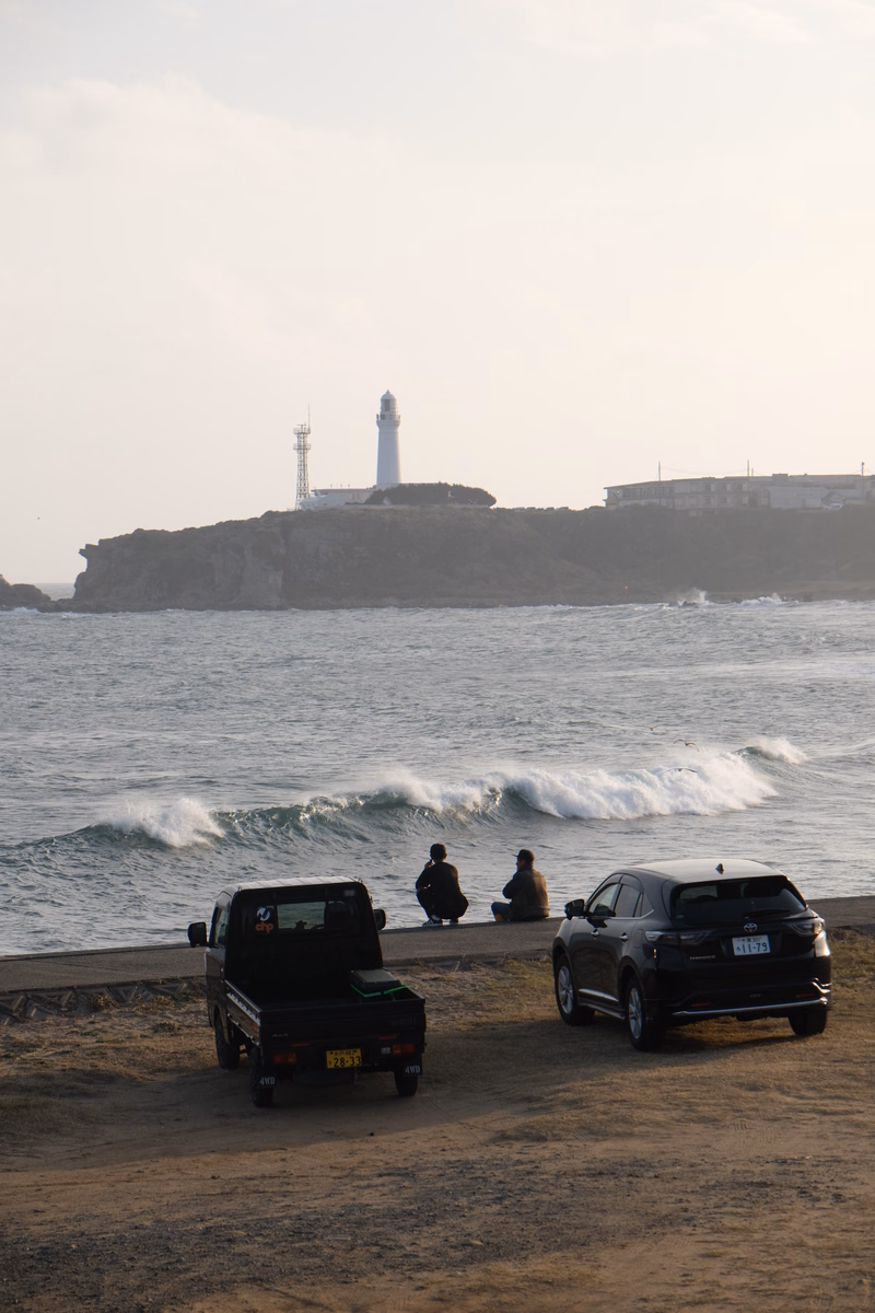 A white lighthouse stands on a rocky coastline overlooking the ocean, with two people and two cars parked on the sandy beach.