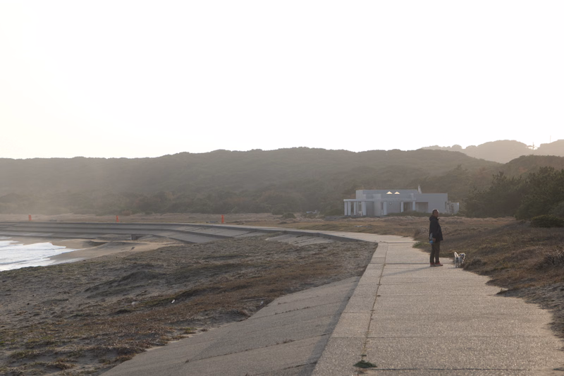 A person walks a dog along a paved path near a white beach hut on a coastal landscape.