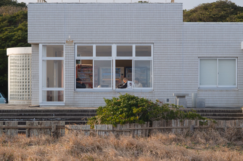 A modern, light-colored tiled shop building with large windows is set against a backdrop of green trees and dry grass.