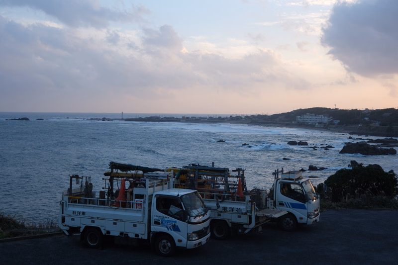 Several white trucks are parked on a paved area overlooking the ocean at sunset near the coastline.