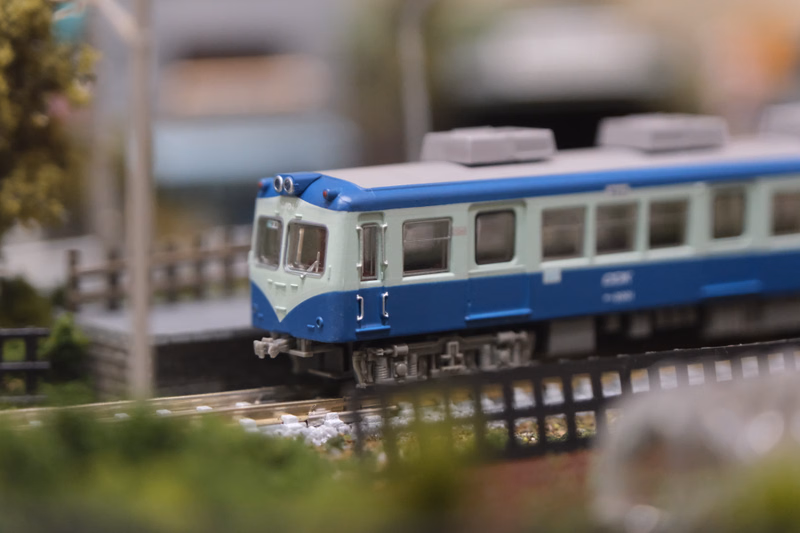 A blue and white model train is running on a miniature railroad track outdoors.