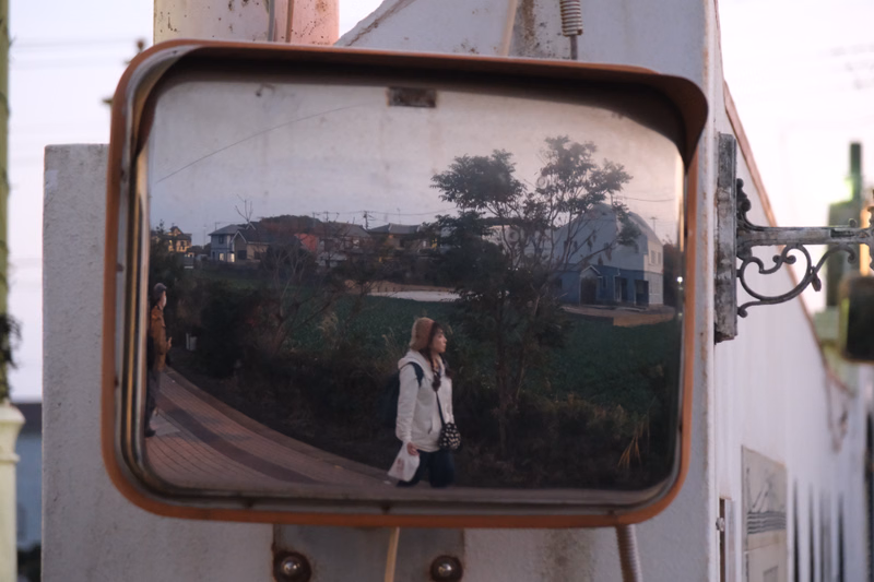 A woman walks down a street reflected in a large, framed mirror mounted on a white structure.