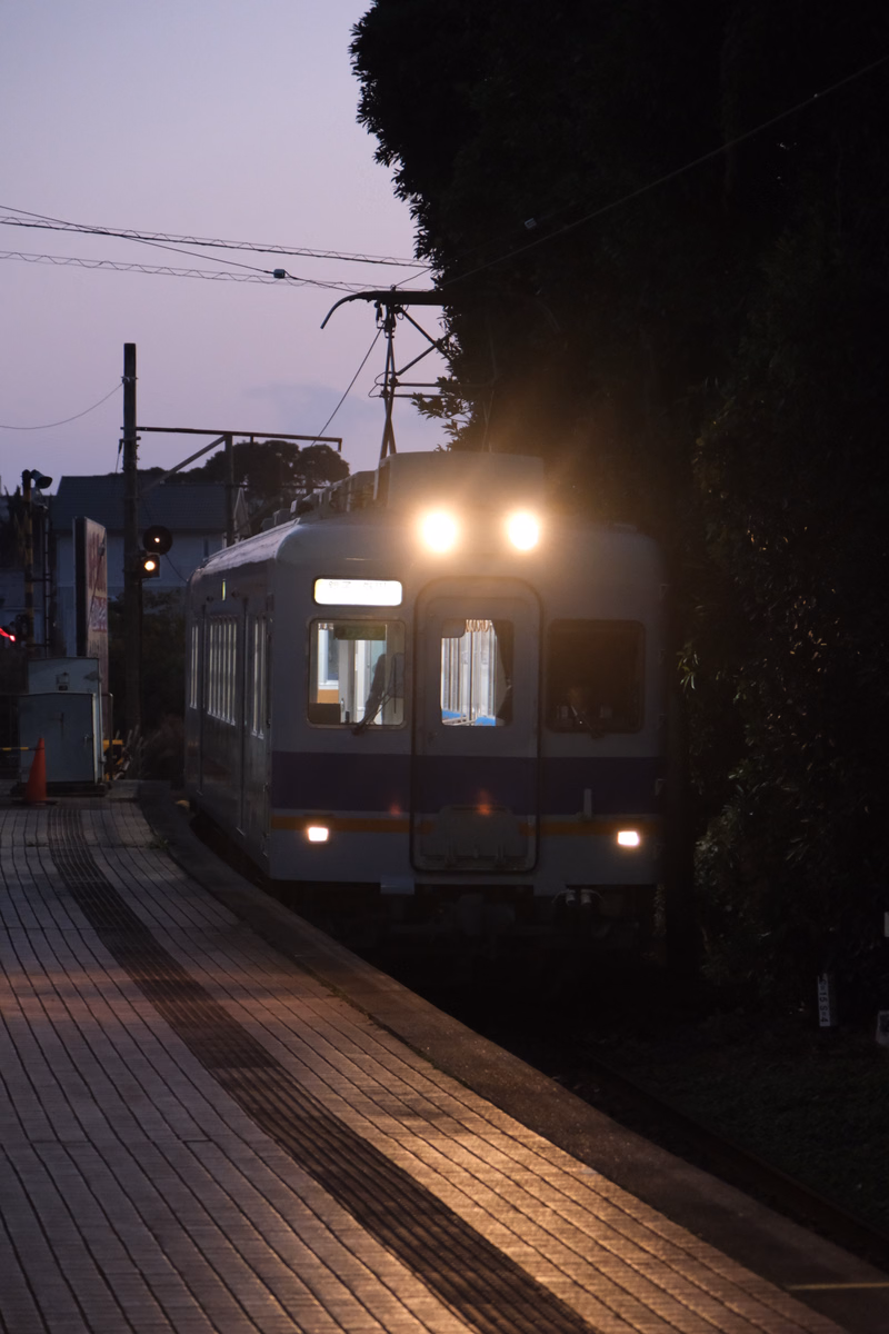 A white and blue train is visible on the tracks near a station platform during the evening.