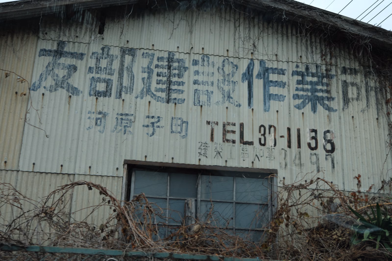 A weathered industrial building with large Japanese characters painted on the side, including "TEL 37-1138".