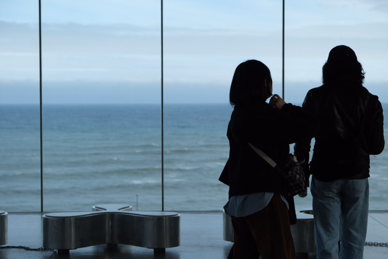 Two people stand looking out at the ocean through a large window overlooking the coast.