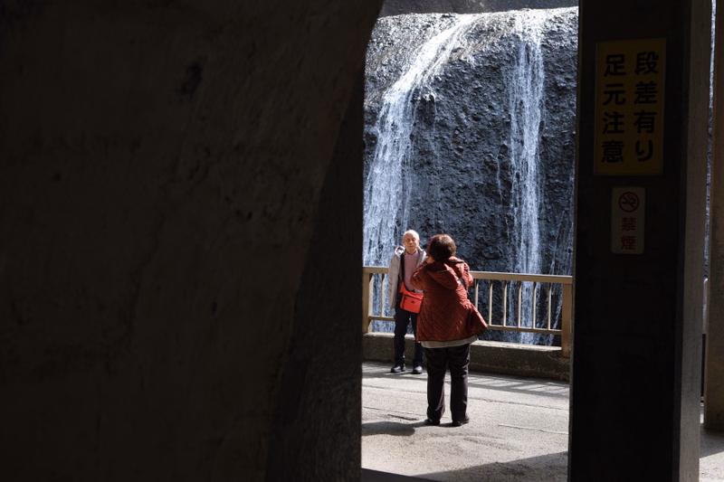 Two people stand near a large waterfall, with a sign nearby that reads "段差有り 足元注意".