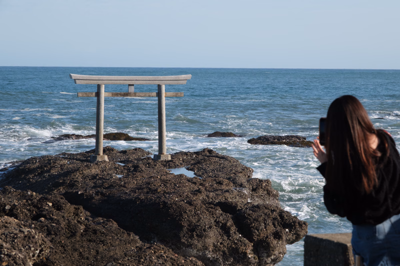 A wooden torii gate stands on dark rocks overlooking the blue ocean while a person takes a photo.