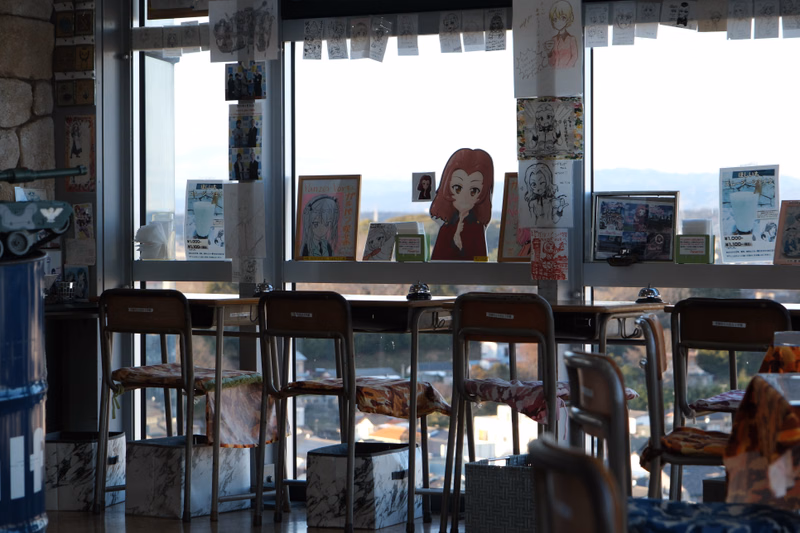 A cafe interior features several barstools facing a large window displaying anime artwork, including a character print of a girl.