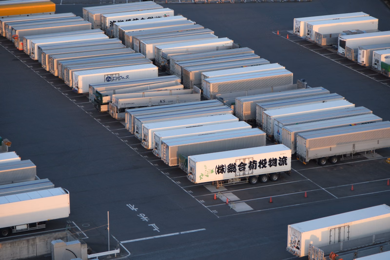 A large outdoor lot filled with numerous stacked shipping containers, some marked with "(株)徳谷輸送".