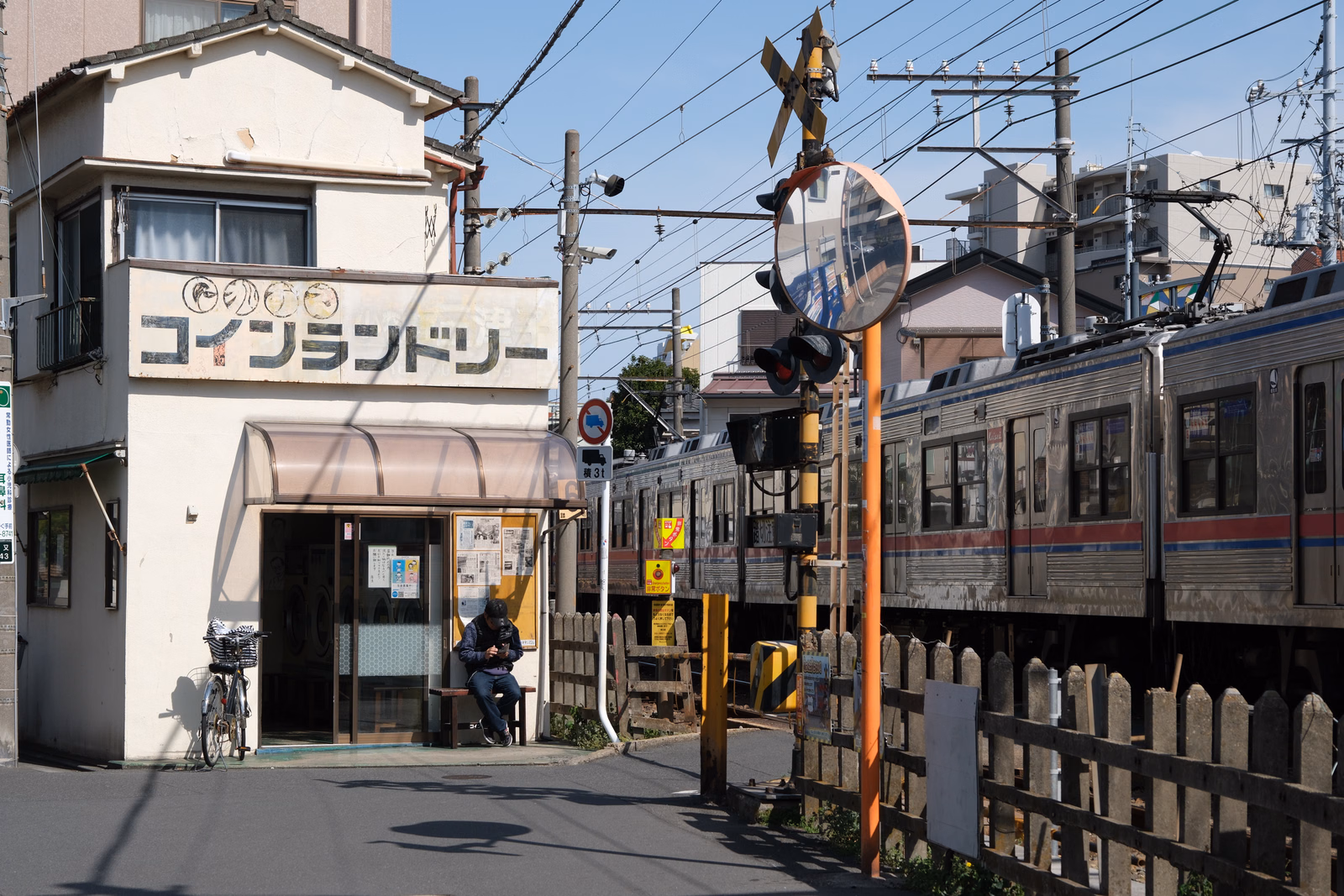 A coin laundry with a sign reading "コインランドリー" is next to a railway track with a train passing under overhead wires.