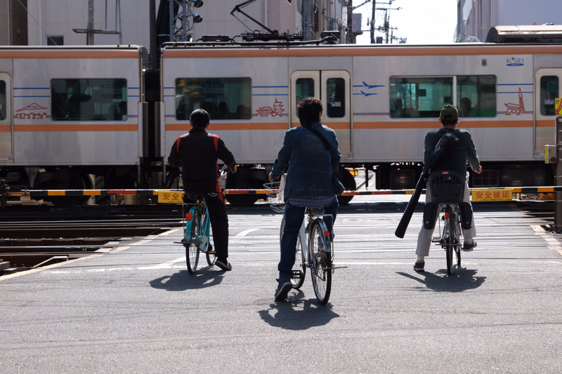 Three cyclists ride on an asphalt street in front of a parked train with "KSEI" visible on the side.
