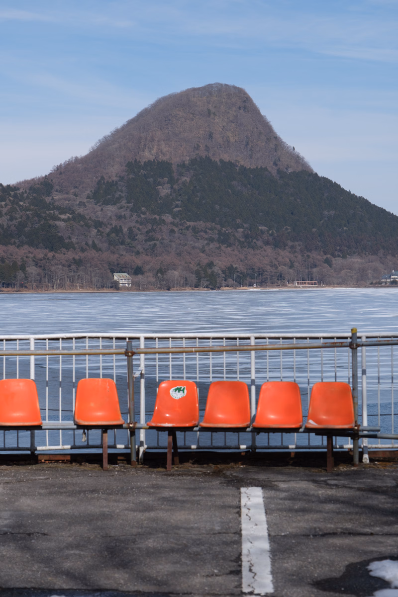 A view of a large, tree-covered mountain rising behind a wide expanse of water, with several orange plastic chairs lined up in the foreground.