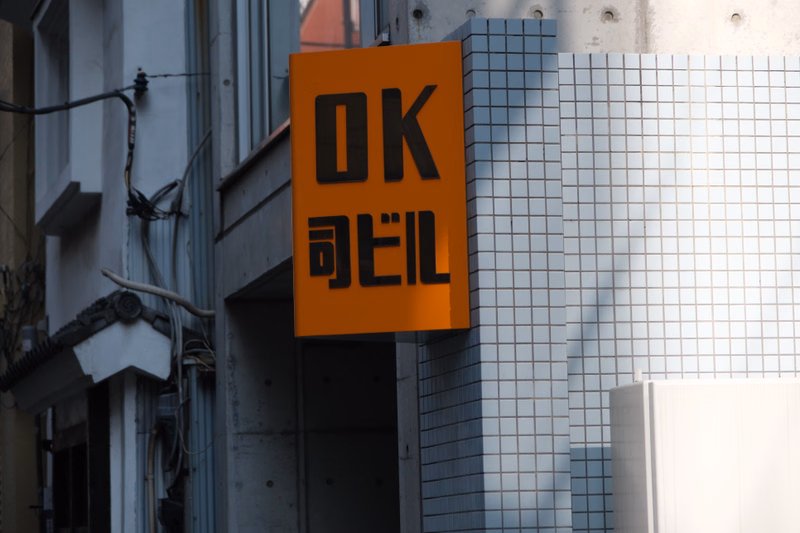 An orange sign displaying "OK 回ピル" is mounted on a modern building facade with a grid pattern.