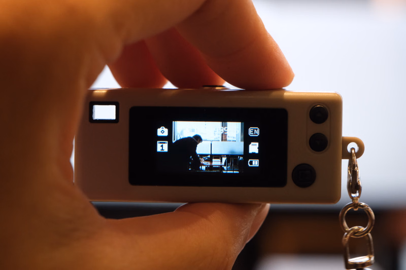 A person holds a small digital camera that is actively recording video, showing a scene with a person inside.
