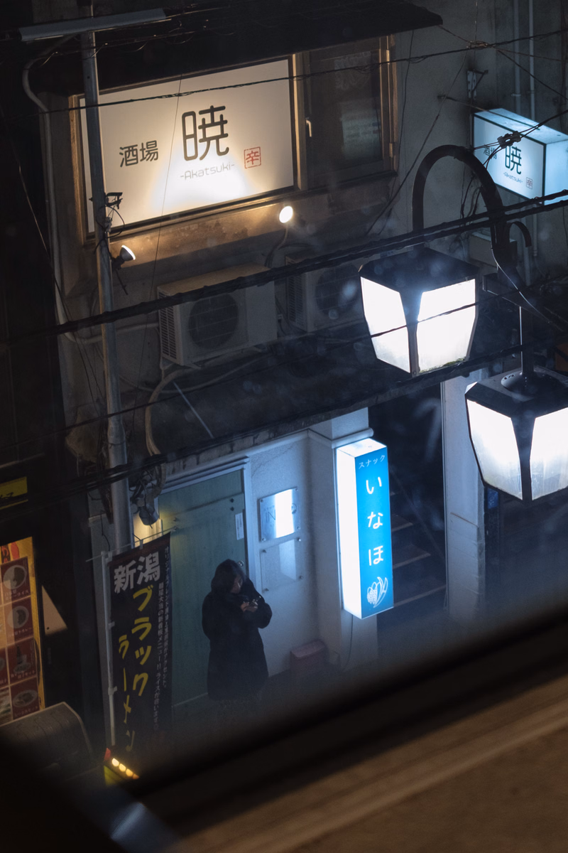 A nighttime street scene featuring illuminated Japanese signs, including one that reads "酒場 暁 -Akatsuki-".