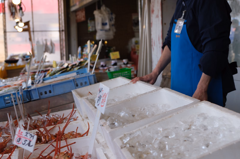 A vendor in a market displays fresh crabs on beds of ice in white containers, with a sign that reads "えび 5元" visible.
