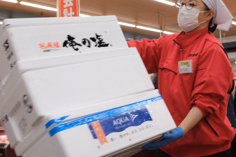 A person wearing a mask holds a stack of white boxes labeled "富沢 竜の塩" and a box with "AQUA" branding.
