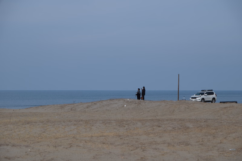 A few people stand on a sandy beach overlooking the ocean, with a white car parked nearby.
