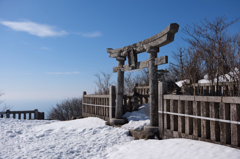 A stone torii gate stands in the snow near a wooden fence overlooking a vast blue sky.