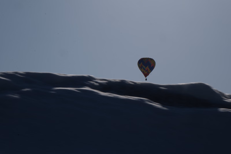 A colorful hot air balloon floats above a snowy, undulating landscape under a clear sky.