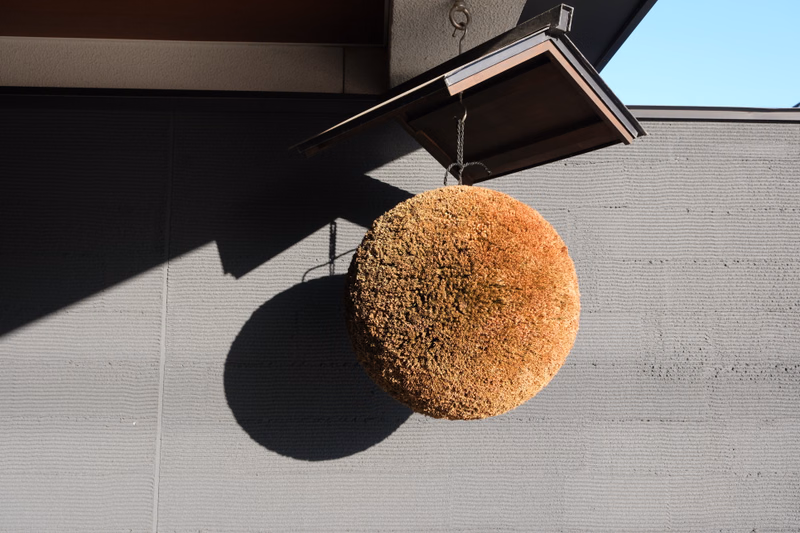 A large, textured, spherical pom-pom decoration hangs from a small wooden structure against a light gray textured wall under bright sunlight.