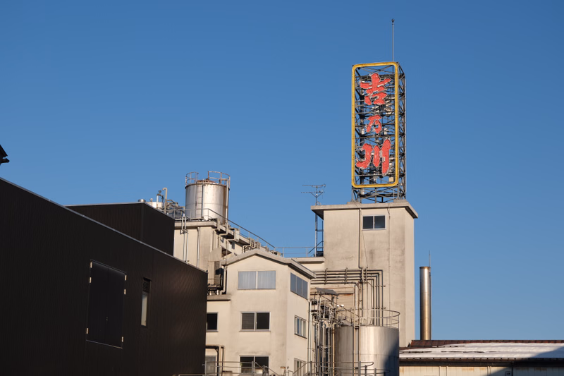 A large, illuminated billboard displaying Japanese characters, "京王", stands above a complex of industrial buildings against a clear blue sky.