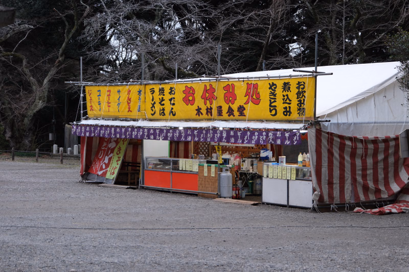 A Japanese food stall with a yellow banner reading "お休みの食堂" is set up outdoors, featuring various food items.
