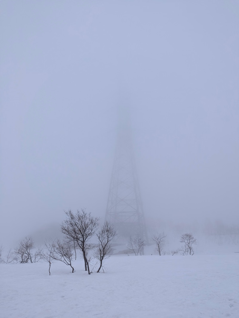 A large radio tower stands in a snowy, foggy field with several bare trees in the foreground.