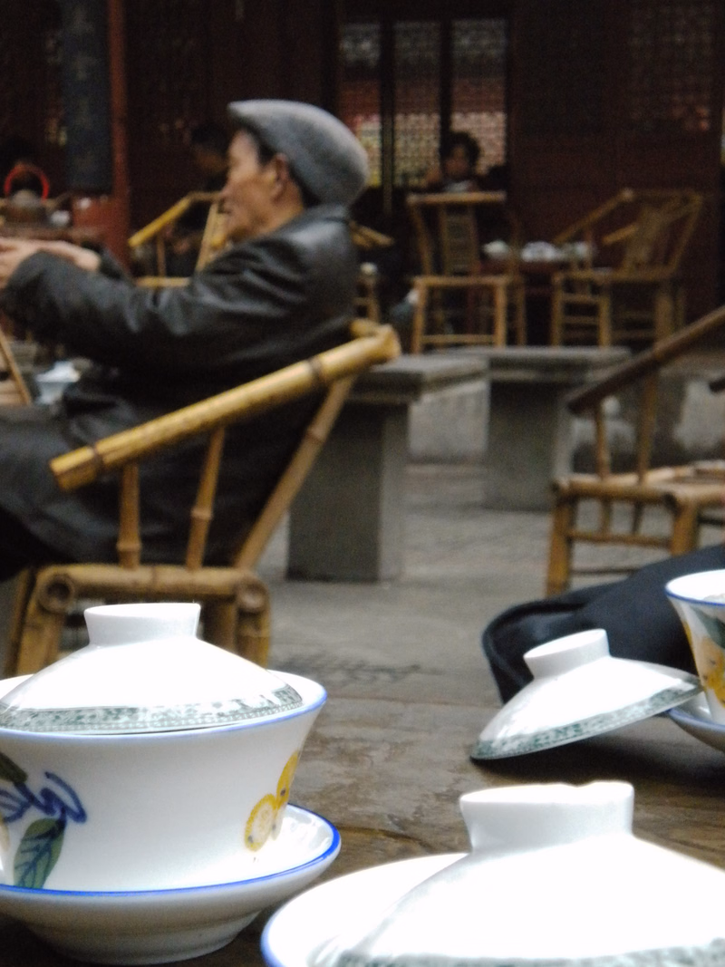 A man in a grey cap sits on a wooden chair outdoors near a table with white ceramic tea cups.