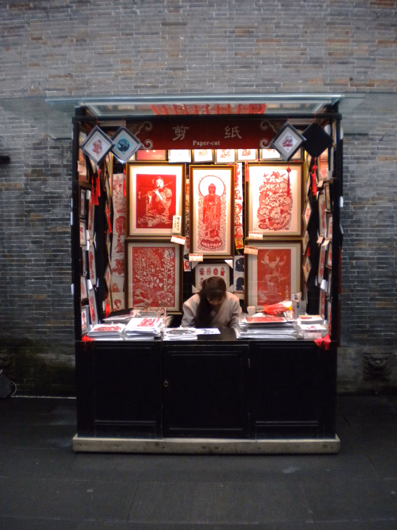 A woman stands behind a kiosk selling traditional Chinese paper art.