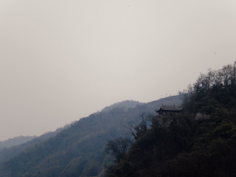 A traditional temple structure is nestled on a densely forested hillside under a hazy sky.