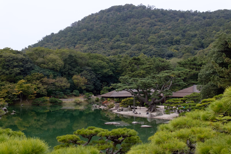 A traditional pavilion sits near a calm pond surrounded by lush green trees and a large forested mountain in the background.
