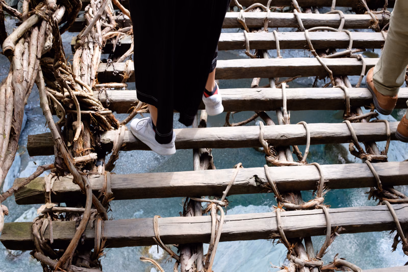 A person wearing dark pants and white sneakers walks across a rustic wooden walkway built over the ocean water.
