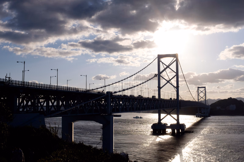 A large suspension bridge spans over a body of water during a bright sunset with dramatic clouds overhead.