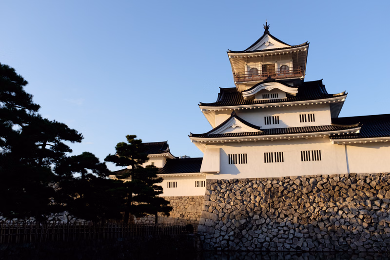A traditional Japanese castle with white walls and dark tiled roofs stands behind a large stone retaining wall under a clear blue sky.