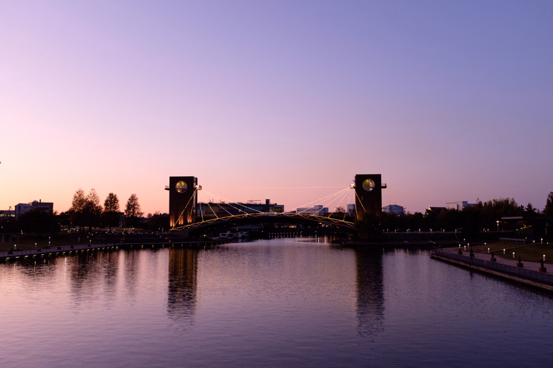 A suspension bridge with illuminated towers spans a wide river during twilight, reflecting the soft colors of the sky.