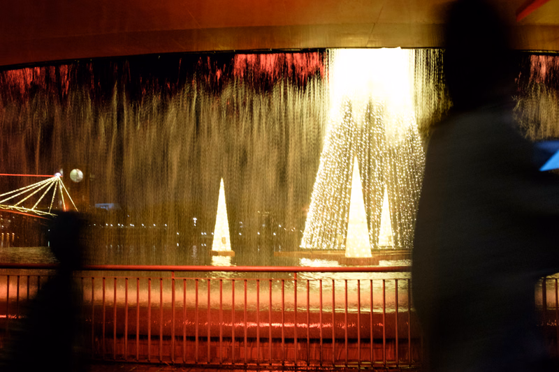 A nighttime view of a large illuminated water fountain with several conical light features, viewed from behind silhouetted people.