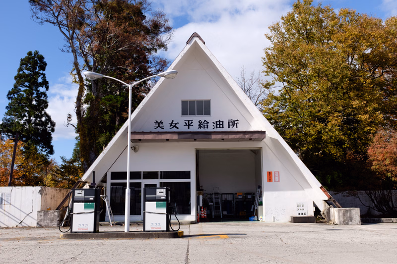 A white, A-frame building with a sign reading "美女平給油所" stands in an outdoor paved area.