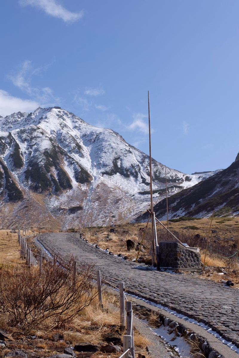 A winding gravel road leads through a mountainous landscape with patches of snow under a clear blue sky.
