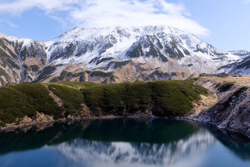 A clear reflection of a snow-capped mountain range is visible in a dark alpine lake under a partly cloudy sky.