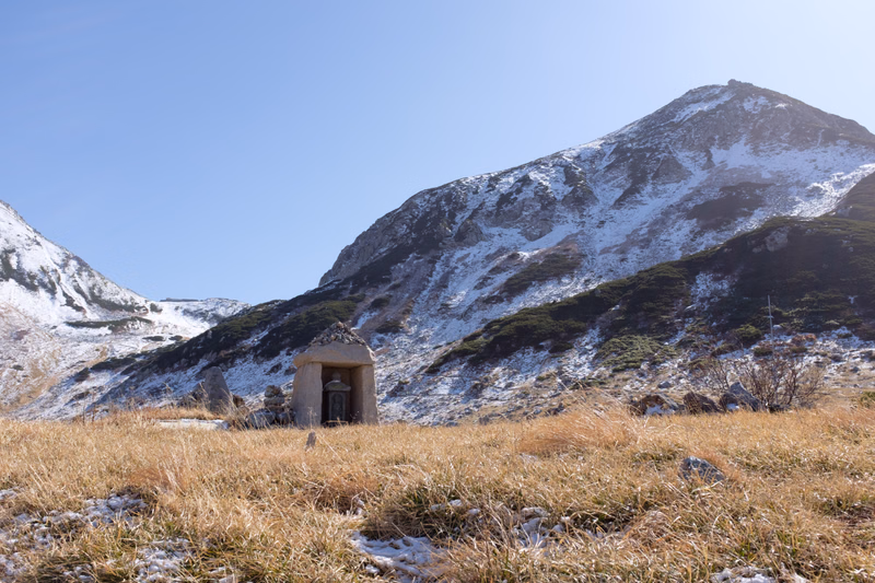 A small stone structure sits in a grassy field with patches of snow, set against a large, snow-dusted mountain under a clear blue sky.