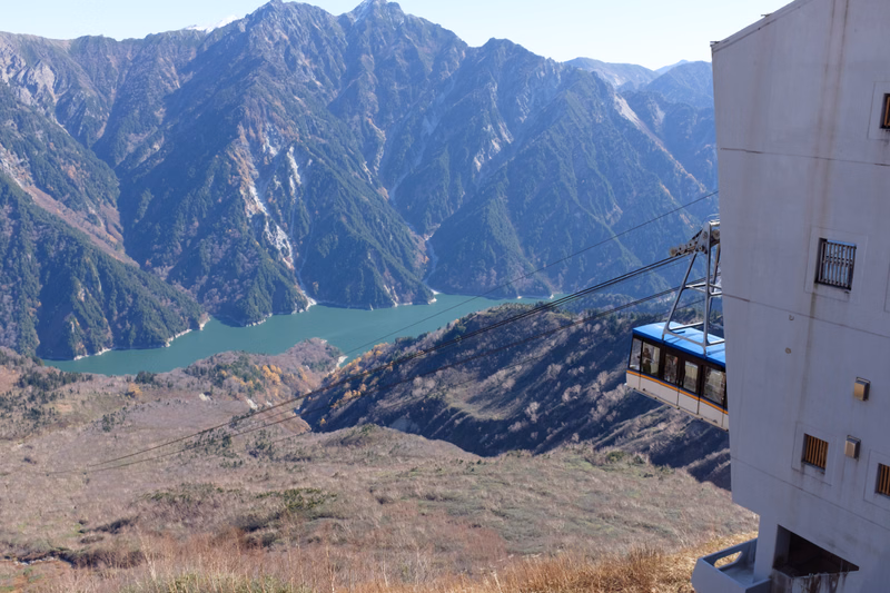 A cable car ascends past a modern building overlooking a large lake surrounded by steep, forested mountains.