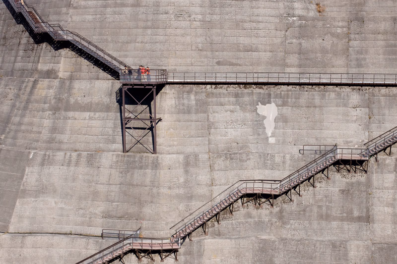 A wide view of concrete steps leading up a large dam structure, featuring metal railings and a small observation platform.