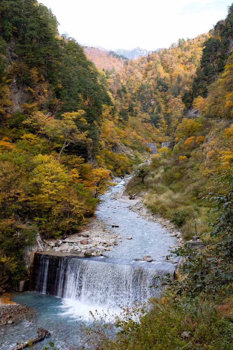 A wide river flows through a forested valley, featuring a small, tiered waterfall cascading into the water below.