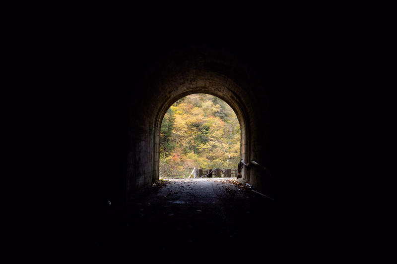 A dark concrete tunnel archway opens up to a bright view of a forest road surrounded by autumn trees.