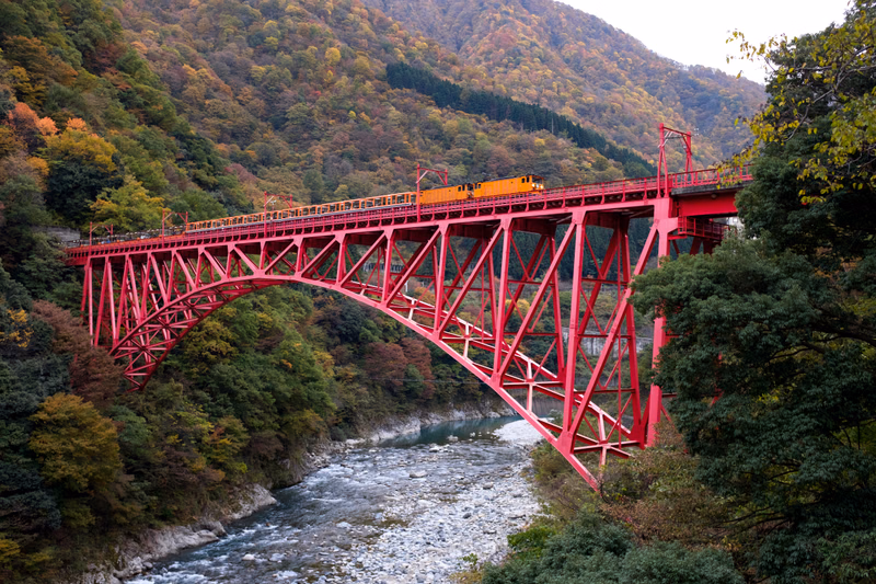 A bright red truss railroad bridge crosses a rocky river through a forested mountain valley, with a train visible on the tracks.