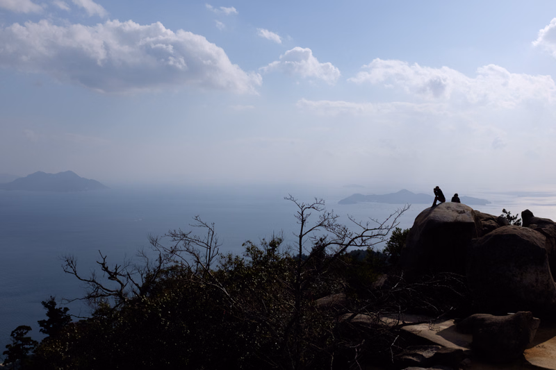 A few people are visible on a rocky outcrop overlooking a vast ocean under a cloudy sky.