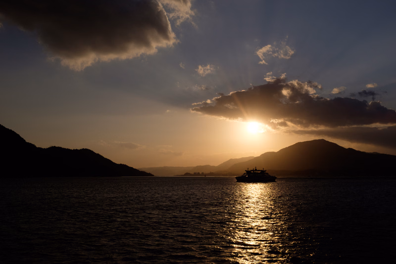 A boat is silhouetted on the water during a bright sunset over mountains.